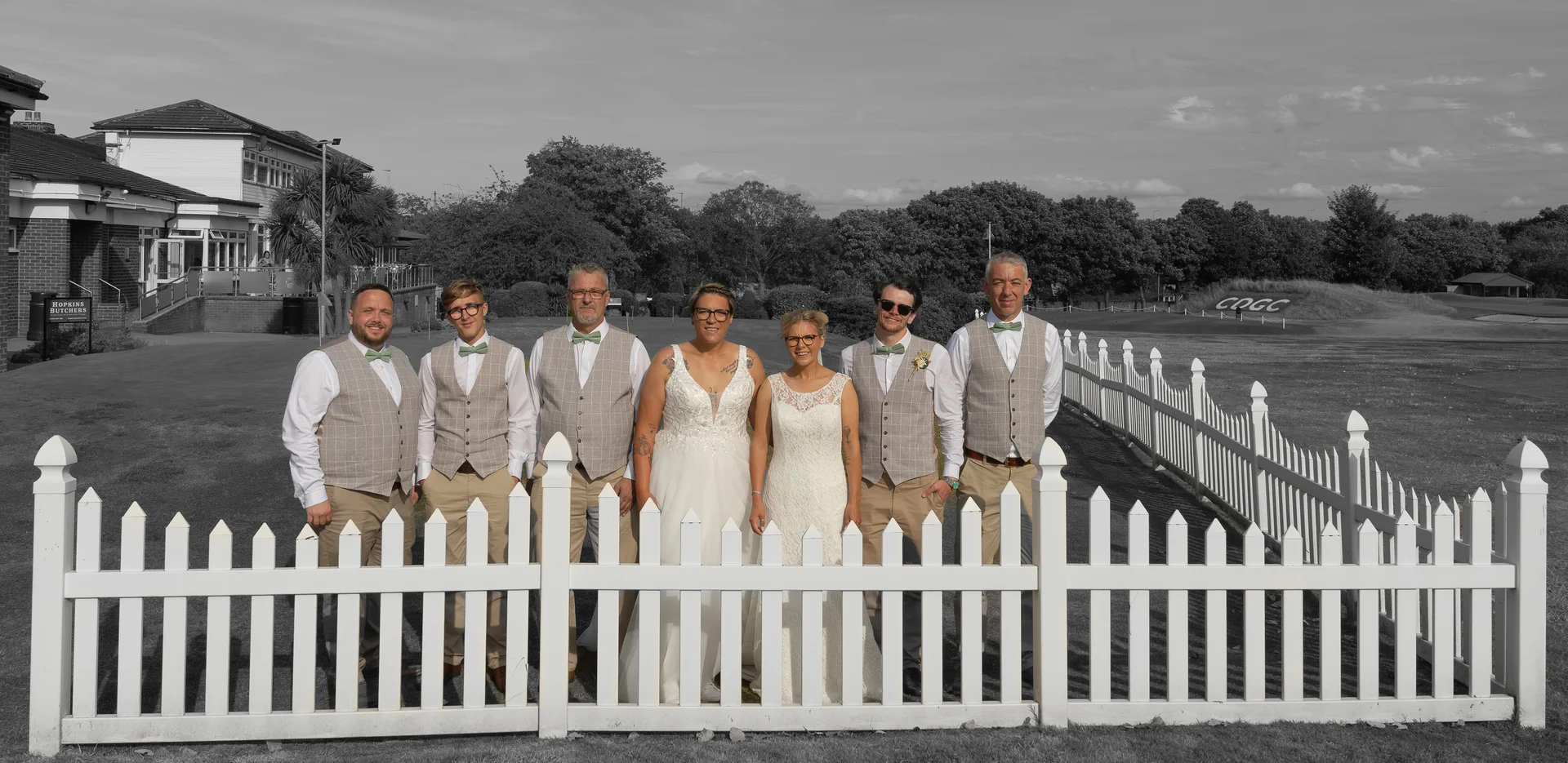 Wedding party gathered behind a white picket fence, with the couple at the centre.